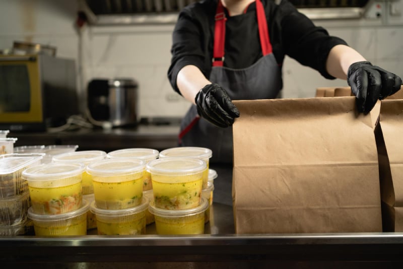 Gloved worker packing takeout containers into a brown paper bag.